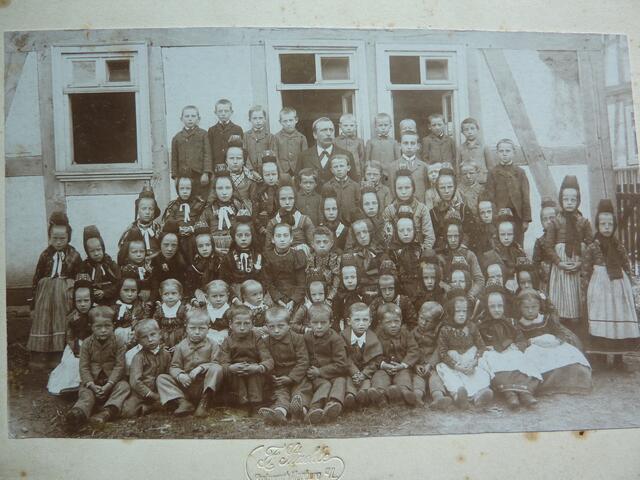 Mehrere Jahrgänge auf dem Klassenfoto mit Lehrer KORNMANN um 1900; vor dem Fachwerk-Schulgebäude.
