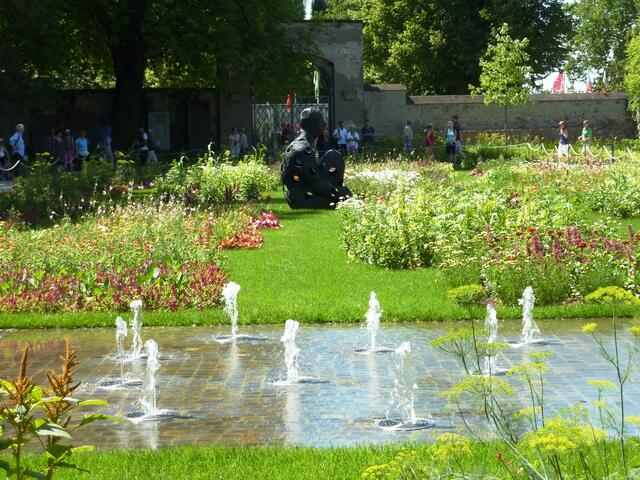 Markante Skulptur inmitten von zarten Blumen und Wasserspielen.