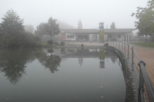 Der Nebel lässt sich alle Zeit, und dem Eingangspavillon des Kurparks bleibt nur ein mattes Spiegelbild im Parksee.