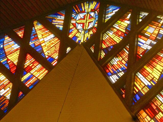 Kirchenfenster der Auferstehungskirche Döhren. Unter dem farbigen Kreuz, auf der Spitze, der hellen Wand, brütet eine Schwalbenfamilie, die uns z. T. lautstark unterhielt