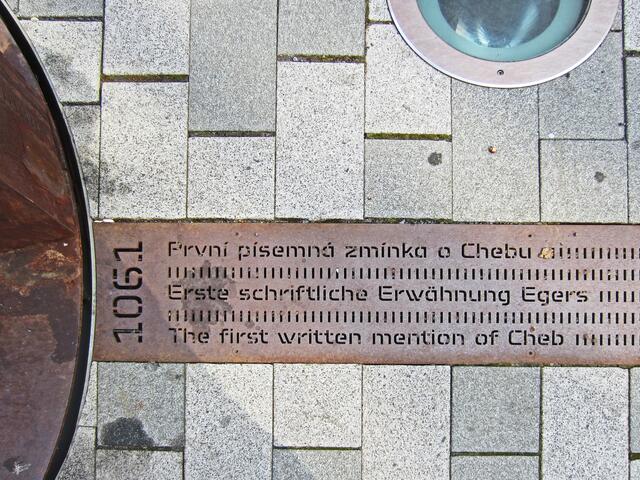 Marktplatz von Cheb (13. Jhdt.).
Denkmal in Form einer Metallsäule.
Von hier aus führt ein Metallband bis zum Bahnhof.
Darauf sind Inschriften aller geschichtlichen Ereignisse der Stadt Eger eingraviert, angefangen von der erstmaligen urkundlichen Erwähnung der Stadt (1061) bis in die Neuzeit.