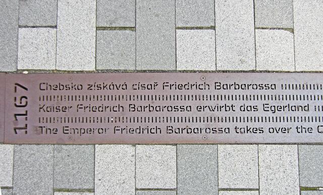 Marktplatz von Cheb (13. Jhdt.).
Denkmal in Form einer Metallsäule.
Von hier aus führt ein Metallband bis zum Bahnhof.
Darauf sind Inschriften aller geschichtlichen Ereignisse der Stadt Eger eingraviert, angefangen von der erstmaligen urkundlichen Erwähnung der Stadt (1061) bis in die Neuzeit.