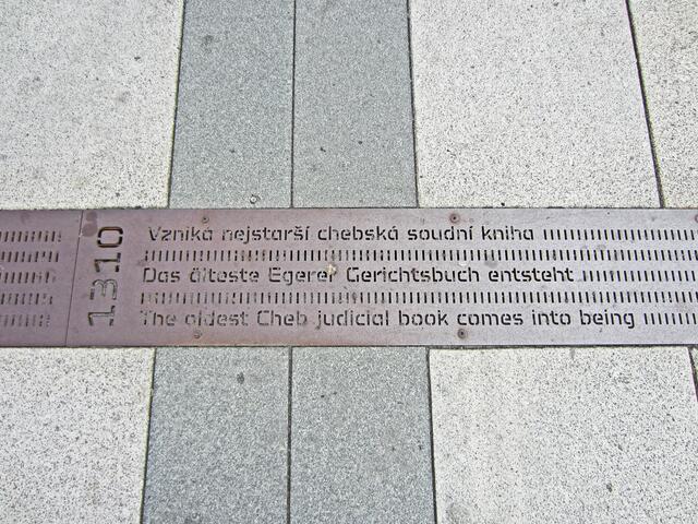 Marktplatz von Cheb (13. Jhdt.).
Denkmal in Form einer Metallsäule.
Von hier aus führt ein Metallband bis zum Bahnhof.
Darauf sind Inschriften aller geschichtlichen Ereignisse der Stadt Eger eingraviert, angefangen von der erstmaligen urkundlichen Erwähnung der Stadt (1061) bis in die Neuzeit.