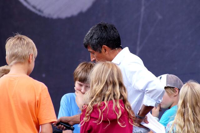 Ranga Yogeshwar inmitten einer Gruppe wissenschaftlich interessierter Kinder, die sich mit großem Interesse an der Lösung einer Aufgabe beteiligen.