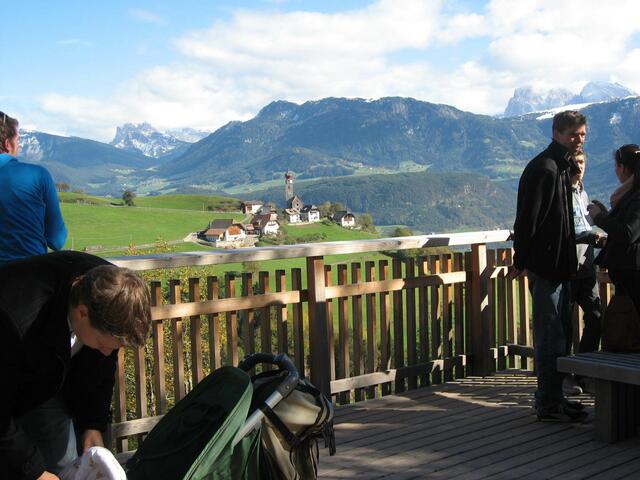 Hier die hölzerne Aussichtskanzel mit Blick hinüber nach Mittelberg zur Kirche St. Nikolaus. Im Hintergrund verschneite Dolomitenberge.