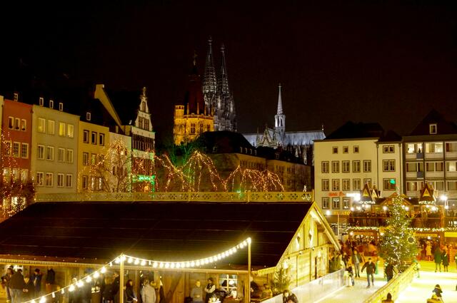 Im Vordergrund sehen wir die in die "Heimat der Heinzel" eingebettete Eisbahn auf dem Heumarkt. Im Hintergrund erscheint im Lichterglanz der Rathausturm und im Schein der nächtlichen Beleuchtung der Kölner Dom.