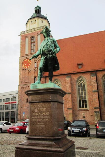 Das Denkmal am Schlossplatz vor der Marienkirche zeigt Leopold, Fürst zu Anhalt, königlich preußischer und Reichs-Generalfeldmarschall (05.07.1676 – 09.04.1747). Der Geburtstag ist hier wohl falsch wiedergegeben, da es der 03.07. war. Den Grund hierfür kenne ich jedoch nicht.