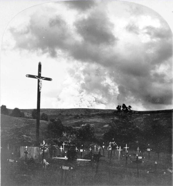 „‘Tis now the very witching time of night: When churchyards yawn, and hell itself breathes out Contagion to this world“ (Es ist jetzt die Zauberzeit der Nacht: Wenn Kirchhöfe gähnen und die Hölle selbst dieser Welt Ansteckung aushaucht) – Friedhof in Saint-Anne auf der Insel Martinique mit einem großen Kruzifix und Kreuzen auf Gräbern. Im Hintergrund steigt dichter Rauch von der Montagne Pelée auf. Fotodruck auf einer Stereo-Karte der Keystone View Company, Meadville, Pennsylvania, 1903. | Foto: Keine bekannten Einschränkungen bei Veröffentlichung. Digitale Sammlung Blazek