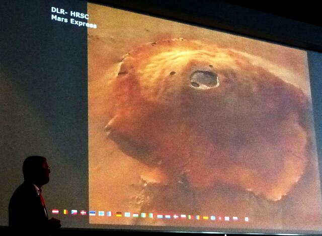 Reiter erläutert, wie ein Marsbesuch ablaufen wird. Er stellt den Olympus Mons vor, den höchsten Berg des Sonnensystems. Er hat die dreifache Höhe des Mount Everests.