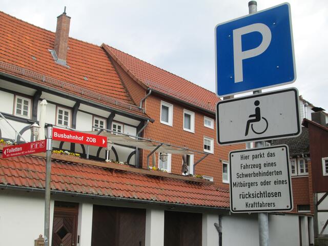 Hinweisschild unter einem Behinderten-Parkplatz hinter dem Rathaus: schwerbehinderter oder rücksichtsloser Kraftfahrer!