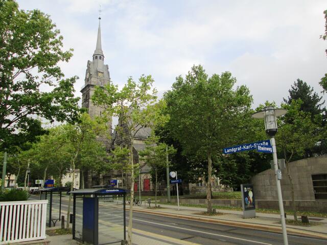 Die Christuskirche der Evangelischen Kirchengemeinde Bad Wilhelmshöhe - eine Ansicht aus der Landgraf-Karl-Straße mit der Haltestelle "Christuskirche" der Tram, die von der Kasseler Verkehrs-Gesellschaft Aktiengesellschaft betrieben wird. Vorne rechts ist eine Außenmauer des Gemeindepavillons zu sehen, der 2001 eingeweiht wurde.