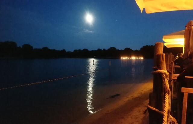 Der Vollmond spiegelt sich im See. Im Hintergrund sind die Fackelschwimmer unterwegs.