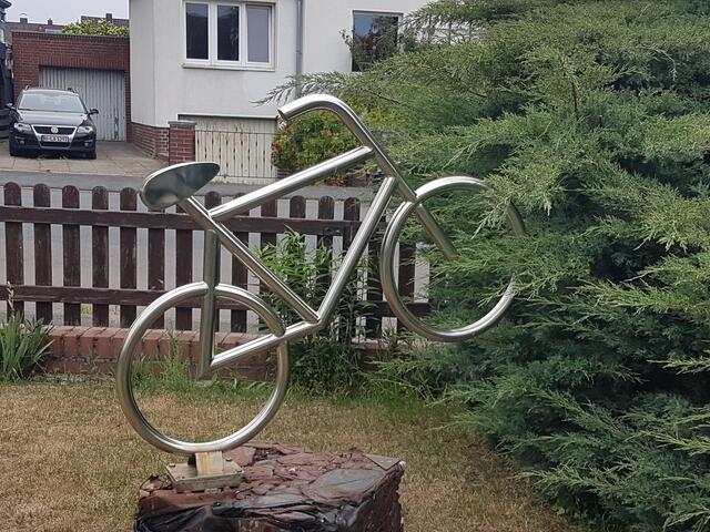 Das silberne Rad aus unvergänglichem, rostfreien Edelstahl steht auf einem Metallblock, entstanden aus einem in einer Schrottpresse zusammengepressten Opel Kadett: Diese Skulptur signalisiert den Sieg des Fahrrades über das Auto.
