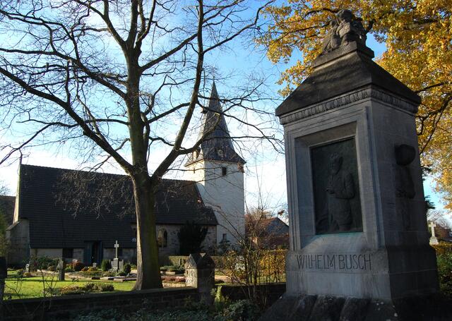 Das Denkmal vor dem Pfarrhaus. Im Hintergrund die 750 Jahre alte St. Nicolai Kirche, in der Wilhelm Busch als Kind sicher so manches Mal die harte Bank drücken musste. Sie wurde aus Feldsteinen und groben Sandstein errichtet.
