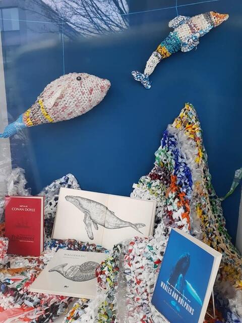 Verschiedene Buchempfehlungen und Illustrationen des Künstlers Paul Katoe liegen am Plastikmeeresgrund, während darüber ein Delfin und ein kleiner Wal schwimmen.