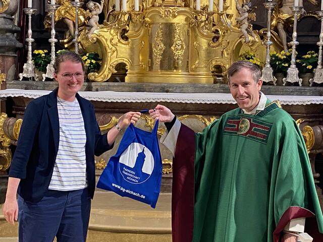 Stadtpfarrer Herbert Gugler überreicht Pastoralreferentin Julia Winter eine prallgefüllte PG-Aichach-Tasche