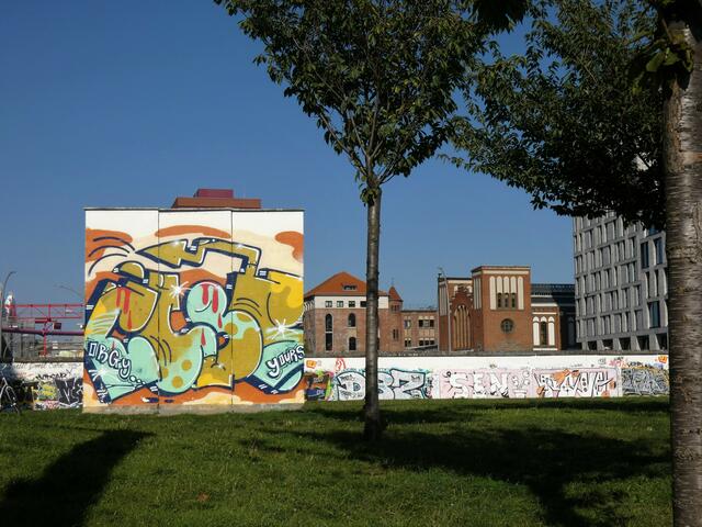 Mauerstück im Park an der Spree. Das rote Backsteingebäude ist der ehemalige Postbahnhof, in dem sich auch der Fritz-Club befindet.