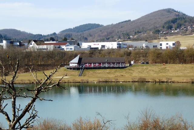 Blick über den See zur Gaststätte Seeblick mit ihrer Terrasse. Oben am Waldrand liegen die Häuser des Ortes Wiesenbach, der Stadtteil von Breidenbach ist.