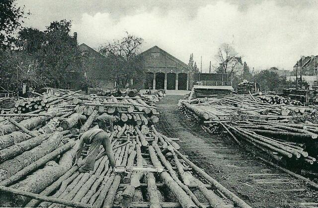 September - Um 1950 geht unser Blick über das Holzlager von ROBERT MOHRMANN (1908-1995) bis zu den Wagenhallen der Straßenbahn. Mit 18 Jahren machte er sich an der SUTELSTRASSE 71 als Stellmacher und Böttcher selbständig. Seine Frau EHLY führte den Betrieb von 1944-1949, während ihr Ehemann ROBERT in russischer Kriegsgefangenschaft war. Auf der anderen Seite der Sutelstraße stehen seit 1897 zwei große Wagenhallen sowie das Kessel- u. Maschinenhaus der Straßenbahn.