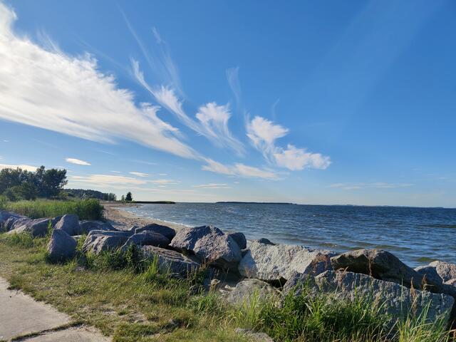 Sandstrand hinter Felsgestein, blaues Meer und blauer Himmel mit eigenwilligen Wolkenbildern.