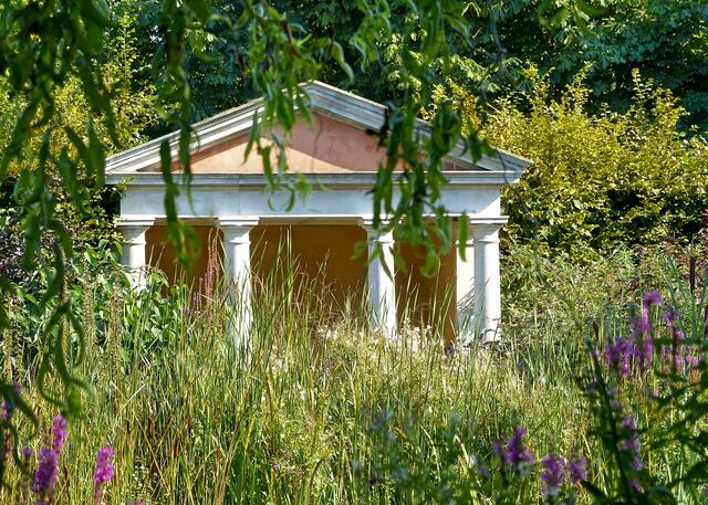 Kleiner griechischer Tempel am Teich umgeben von üppiger Vegetation.