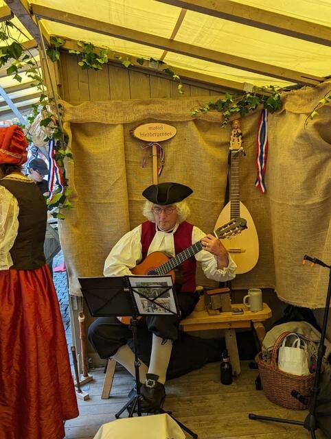 Musikalischer Leiter der "Musici Friedbergenses",  Josef Ditchen, spielt während in der Knopfmanufaktur gearbeitet wird Stücke aus dem 18. Jahrhundert. Seit Januar bereitete sich die Musikgruppe unter seiner Leitung wöchentlich auf die Friedberger Zeit vor, um Stücke von Leopold Mozart, Wolfgang Amadeus Mozart und Valentin Rathgeber und weitere im Quartett vorzutragen. Sein Hauptinstrument, den Kontrabass musste er wegen der hohen Temperaturen als Ausstellungsstück in der Bahnhofstr. 6, "Beim Henggi"  zurück lassen. Seine Gitarrenlaute hatte er aber dabei und diese schmückte den Stand der Knopfmanufaktur. Sein Solospiel auf der Gitarre umrahmte die Arbeit der Knopfmanufaktur.