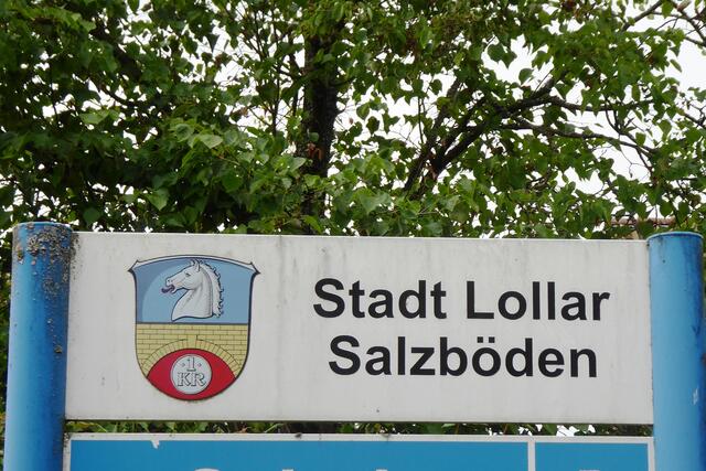 Auf einem innerörtlichen Hinweisschild befindet sich links oben das Wappen der Stadt Lollar. In der oberen Hälfte des waagerecht geteilten Schilds befindet sich in Blau ein silberner, rotbezungter und nach rechts gewendeter Pferdekopf. Unten steht eine goldene Brücke über einem roten Feld, das   mit einer silbernen 1-Kreuzer-Münze belegt ist. 
Hintergrund für die Wappengestaltung ist ein wirtschaftlich wichtiges Ereignis von 1711. Damals wurde ein Brückenzoll über die Lumda genehmigt. Ein Kreuzer wurde für jedes Pferd bei der Überquerung fällig. 
