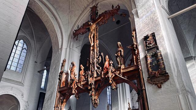 Auf der hohen Tragekonstruktion erhebt sich das spätgotische, reich verzierte Kreuz bis in das Gewölbe. Darunter stehen seitlich Adam und Eva, dann überlebensgroß Maria und Johannes, und unmittelbar neben dem Kreuz knien Maria Magdalena und der Bischof Albert II. Krummendiek, der den Bildschnitzer Bernt Notke (um 1440 bis 1509) beauftragt hatte, dieses Werk anzufertigen. Foto: Helmut Kuzina