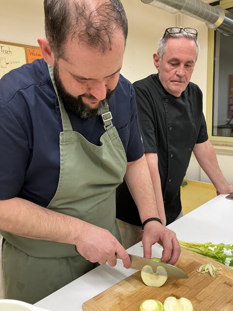 Johannes Steichele schneidet hier die hauchdünnen Apfelscheiben, die dann bis zum Anrichten vakuum verpackt werden. Ganz wichtig dabei ist ein super scharfes Messer. Stefan Zink beobachtet den Profi, wie dieser das macht. 