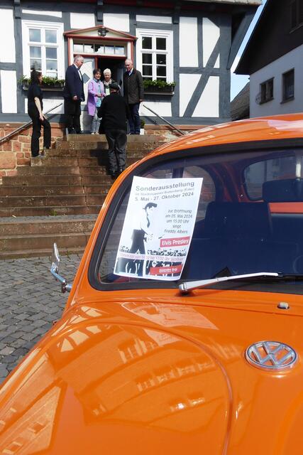 Das Hinweisplakat zur Ausstellungseröffnung auf einem orangen VW-Käfer.