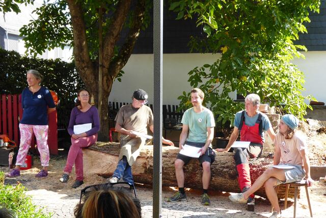 Neben Marlit Peikert vom Kunstverein Battenberg sitzen auf dem Baumstamm die fünf teilnehmenden Künstler: Beate Debus, Matthias Heß, Leon Fiand, Ernst Groß und Ignaz Huber.