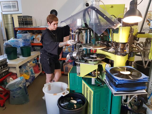 Eine Plattenpresse in betrieb. Große Aufmerksamkeit und Fachkenntnis ist hier erforderlich, dass auch jede Schallplattenpressung den Qualitätsansprüchen genügt.