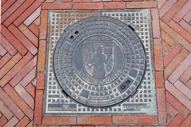 Kanaldeckel mit dem Wappen der Stadt Bremervörde, das auf rotem Grund ein in Silber gehaltenes Bildnis des heiligen Liborius mit Heiligenschein zeigt, der in der Rechten einen silbernen Schlüssel, in der Linken ein offenes Buch hält.