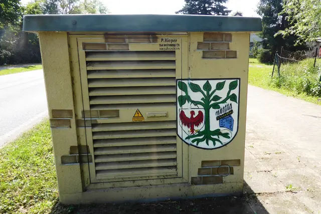 Seitenansicht des Transformatorenhauses in der Berliner Straße mit dem Wappen von Liebenwalde. Das Wappen zeigt: "In Silber eine bewurzelte grüne Linde, beseitet rechts von einem silbernen, an einem Ast hängenden Dreieckschild, belegt mit einem gold-bewehrten roten Adler, und links von einem Topfhelm mit einem schwarzen Flug, belegt mit gestürzten goldenen Lindenblättern."