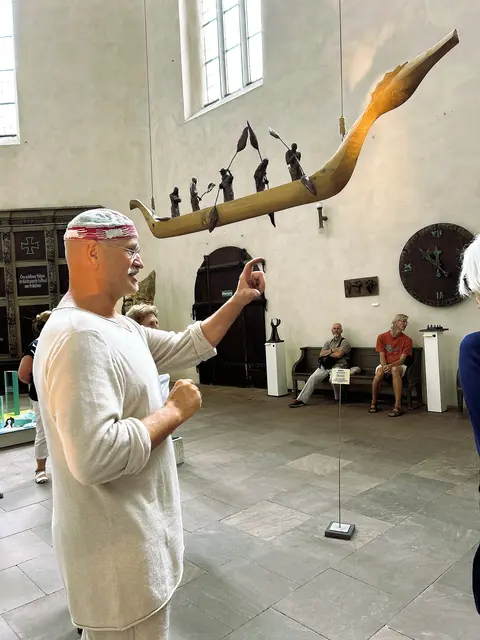 Roland LIndner während einer Führung am 16.08.2025: 
Das Boot mit den Insassen verkörpert die fünf Hauptreligionen. 
Das Kunstwerk ist verkauft. Es darf als Dauerleihgabe in der Kirche verbleiben. 
