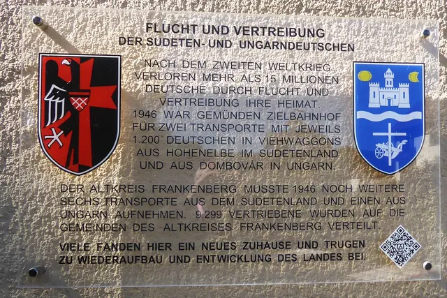 Am 30.07.2018 wurde an der Außenmauer eine Gedenktafel enthüllt. Diese erinnert an die beiden Transporte der Heimatvertriebenen mit jeweils 1200 Personen aus Hohenelbe, Sudetenland, und Dómbovár, Ungarn. Links das Wappen der Sudetendeutschen, rechts das der Ungarndeutschen. Die Burg Buda mit offenem Tor wird von Sonne und Halbmond begleitet, was an die Abwehr der Osmanen erinnert. Die Donau teilt den Schild. Darunter zeigt der  Pflug mit dem christlichen Kreuz die Schaffenskraft und den Glauben der deutschen Siedler.