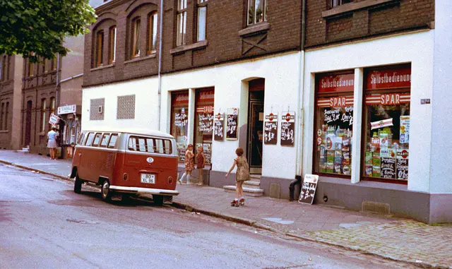 Das Foto zeigt den ehemaligen Lebensmittelladen meiner Eltern und eine Trinkhalle im Ruhrgebiet, in Bochum-Linden, in der Welperstraße, etwa 1966. Damals gab es noch zahlreiche Einzelhandelsgeschäfte, die aber später fast alle verschwanden als die großen Supermärkte aufkamen. Rollschuhlaufen war in diesen Jahren bei den Kindern sehr beliebt, vor allem die mit den Eisenrollen. Kamera: Zeiss-Ikon Contarex | Foto: Volkmar Brockhaus
