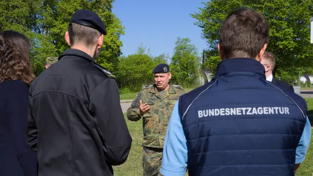 Ein Soldat in Tarnkleidung spricht im Freien zu einer Gruppe. Ein Mann im Vordergrund trägt eine blaue Weste mit Aufschrift Bundesnetzagentur.