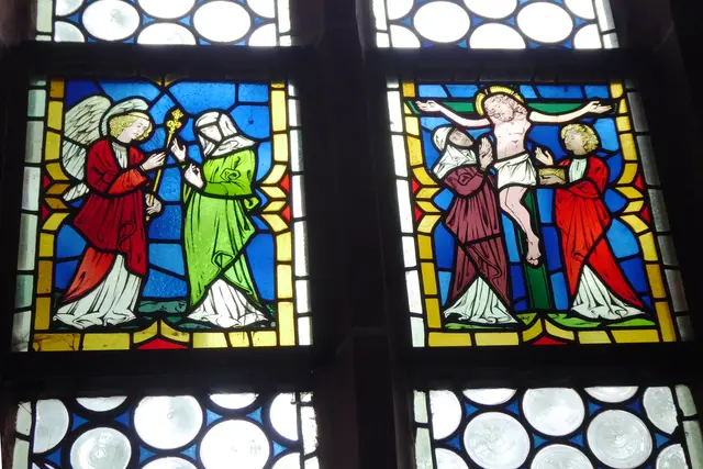 Das Kirchenfenster im Chorraum. Die farbig gestalteten Felder zeigen links die Verkündigung durch den Engel Gabriel, dass die Jungfrau Maria den Sohn Gottes vom Heiligen Geist empfangen und ihn gebären werde, rechts die Kreuzigung Jesu.
