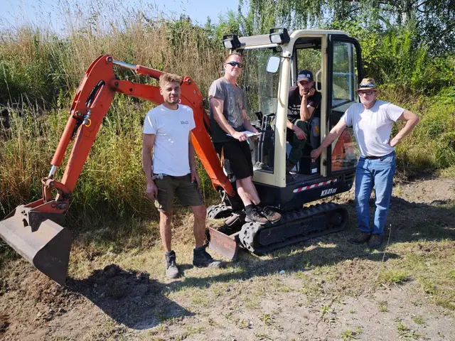Vier Personen stehen an einem Minibagger auf einer Baustelle und arbeiten an der Parkplatzfläche.