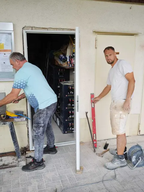 Zwei Personen arbeiten am Eingang des Vereinsheims: Links steht Sven Thymian an einem Arbeitstisch, rechts richtet Jonathan Krause eine neue Feuerschutztür aus.