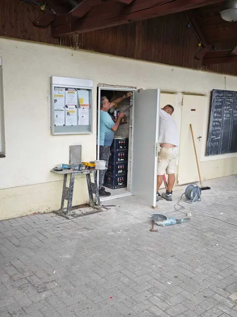 Zwei Personen arbeiten an einem geöffneten Technikschrank am Vereinsheim, während Werkzeuge und Materialien bereitliegen.
