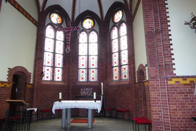 Vor dem Altarraum hängt ein Kreuz aus roten Glasbruchsteinen. In der Mitte der drei kreisförmigen Glasfenster ist eine Herz-Jesu-Darstellung zu sehen, rechts als Patron Deutschlands der Erzengel Michael und links die Heilige Barabara, die Patronin der Marineartillerie.