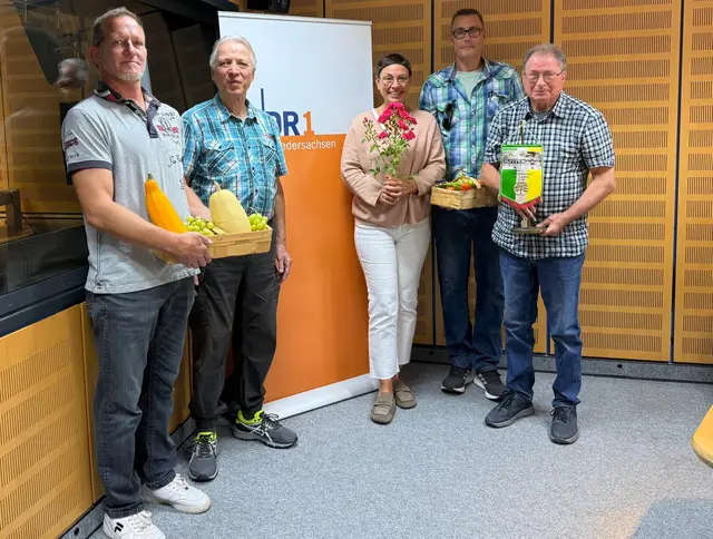 Von links nach rechts stehen Markus Schmidt, Andreas Schmitt, Moderatorin Martina Gilica, Daniel Jansen und Eugenio „Gino“ Maiolo vor einem NDR‑1‑Niedersachsen‑Banner. Sie halten Körbe mit Obst und Gemüse sowie einen Rosenstrauß. Das Bild zeigt die Studio‑Gäste des KGV Dornröschen Lehrte im Landesfunkhaus Hannover.